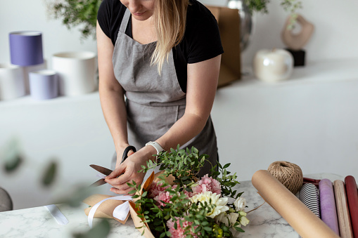 Portrait of female florist in apron arranging fresh flowers for bouquet in the flower shop, using roses, hydrangea, peonies, then wrapping it into color paper and finishing with ribbon