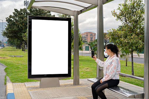 A Latina woman takes a photo with her smartphone of the bus stop billboard.