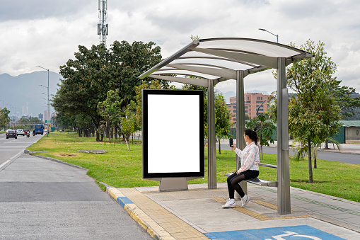 A Latina woman takes a photo with her smartphone of the bus stop billboard.