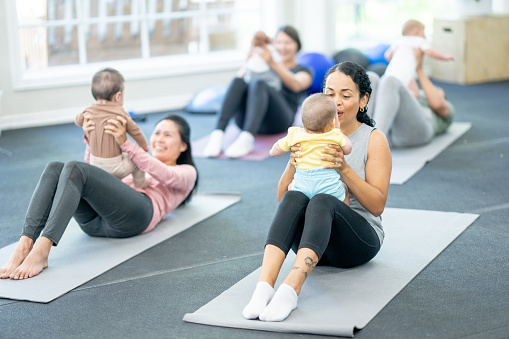A small group of new parents sit on yoga mats in a studio as they hold their babies and participate in a Parent & Baby fitness class.  They are each dressed comfortably and appear to be having fun.