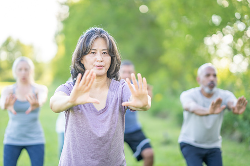 A small group of mature adults practice Tai Chi outdoors together on a warm summer day.  They are each dressed comfortably and are smiling as they take in the fresh air.