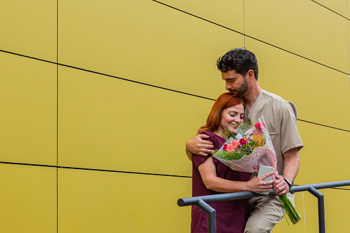 Latin heterosexual couple with an average age of 25 years, a red-haired woman and a man with a black beard, doctors by profession, together dressed in their work uniforms in the clinic. They walk to work while he surprises her with a beautiful bouquet of flowers.