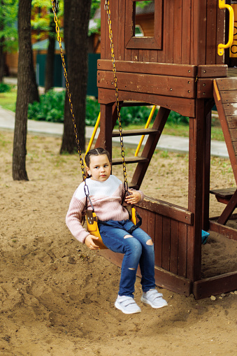 Cute little girl teetering on yellow swing in playground in wooden game complex. Summer holidays in camp, tourist center. Walking and playing outdoors, sport activity and healthy lifestyle.