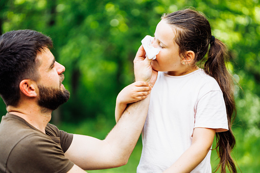 Side view of middle-aged bearded handsome man father wearing brown T-shirt, wiping daughters nose with white handkerchief during walking in summer forest. Family, love, care, flu, sneezing, cold.