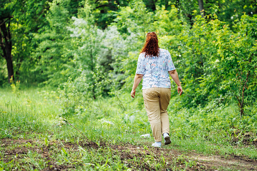 Back view of young overweight woman with long curly red hair in ponytail, wearing blue blouse with floral pattern, beige trousers walking on path in park forest among green trees. Summer, nature.