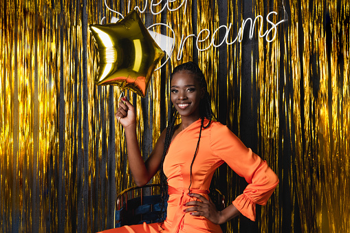 Smiling, merry young african american woman with dreadlocks in orange dress hold festive golden star balloon, look at camera against lametta background. Sweet dreams. Holiday celebration, party night