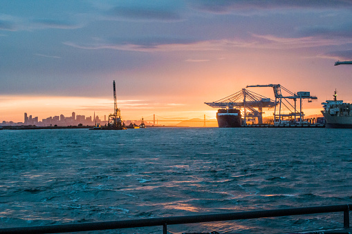 San Francisco Bay Cargo Ship under the Cranes in Silhouette at sunset, futurist clouds with a blue and orange cast