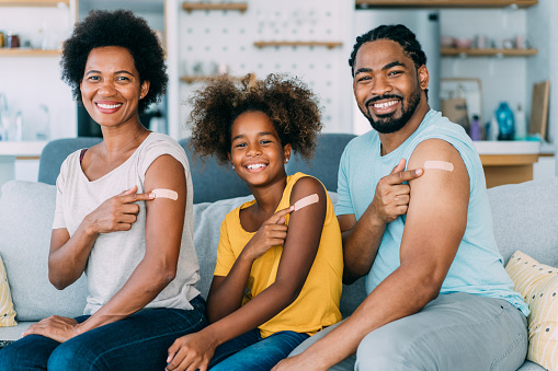 Shot of a smiling family pointing at their arms with a bandage after receiving COVID-19 vaccine. African-american family showing their shoulders after getting coronavirus vaccine. Portrait of father, mother and daughter with adhesive bandages after covid-19 vaccination.