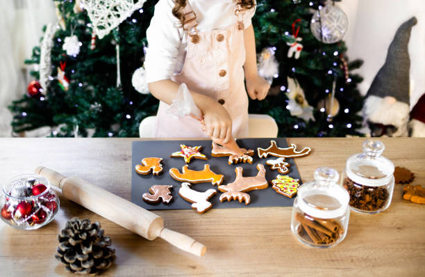 happy little girl coloring christmas gingerbread cookies at home. christmas decoration in the kitchen. fir tree with fairy lights. the concept of the new year and christmas - gift orange green package imagens e fotografias de stock