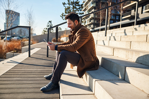 Handsome man wearing coat during sunny autumn sitting with tablet on stairs outside in a city