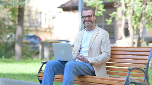 istock Middle Aged Man Smiling at Camera while Working on Laptop Outdoor 1679714217