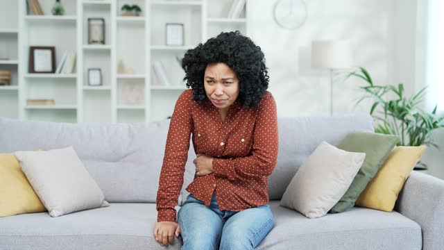 Young african american female suffering from abdominal pain sitting on sofa in room at home.