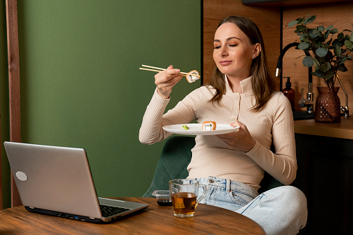 Beautiful young brunette woman dressed in a beige sweatshirt eats sushi rolls with chopsticks in the kitchen.