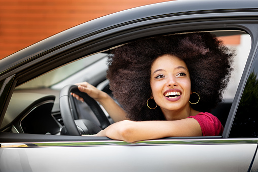 Smiling young Moroccan female with Afro hairstyle looking away while sitting in driver seat of parked car in daylight with hand on steering wheel and elbow on front door window