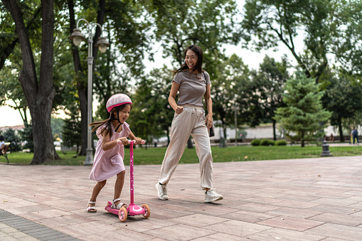 Mother teaching daughter to ride scooter in the public park