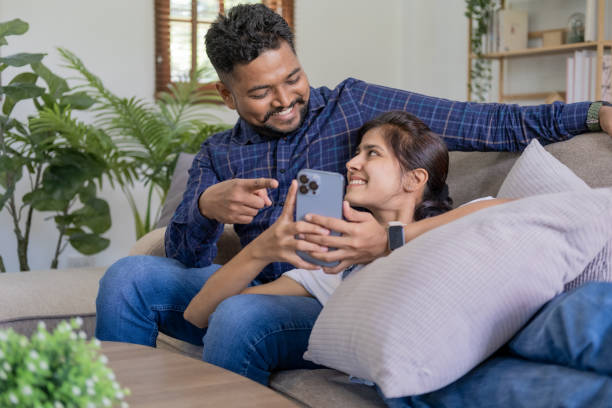 istock Smiling couple embracing while looking at mobile phone, Loving couple relaxing on sofa 1679895071