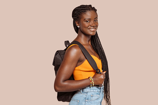 Young african woman with braids, wears yellow top and backpack smiles broadly. Happy smiling black female student at studio isolated over beige background.