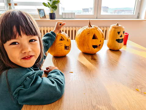 Portrait of a little girl sitting at the kitchen table, holding carved out pumpkins for Halloween.
