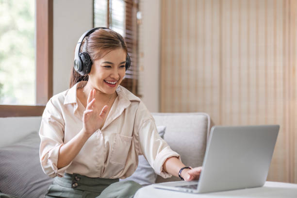 istock Smiling young asian woman using laptop web camera while wearing headphones and sitting on the rug beside to the sofa at homes 1679941744