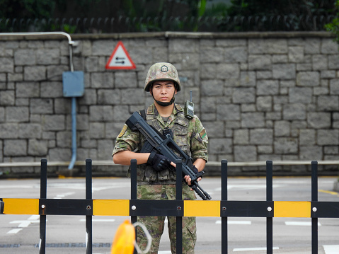 A soldier of the Chinese People's Liberation Army (PLA) guarding the main gate to the PLA Hong Kong Garrison Building in Admiralty, Hong Kong. His gun has a fixed bayonet.  An orange road works light in the foreground marks traffic cones placed in the middle of Lung Wo Road to block motorists turning in front of the entrance.  This image was taken on an overcast afternoon on 14 September 2023.