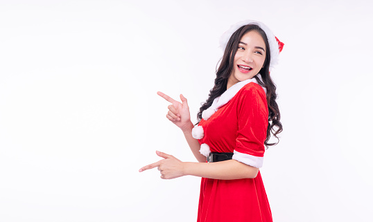 Cheerful surprise asian woman wear Santa Claus dress with Christmas hat use finger pointing present copy space standing on white background. Excited young girl Santa Claus dress merry Christmas winter