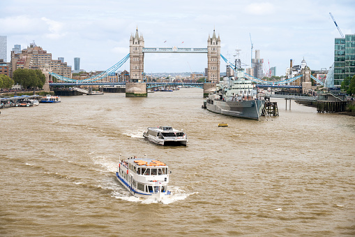 Cityscape of London, United Kingdom. Thames river with floating boats, moored war ship, Tower Bridge and other buildings