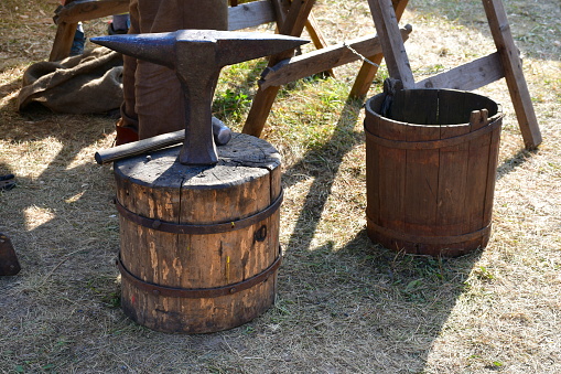 A close up on a big, massive anvil used by a medieval blacksmith located on a big wooden log next to some other equipment and a cloth tent seen in summer during a historic fair or festival in Poland
