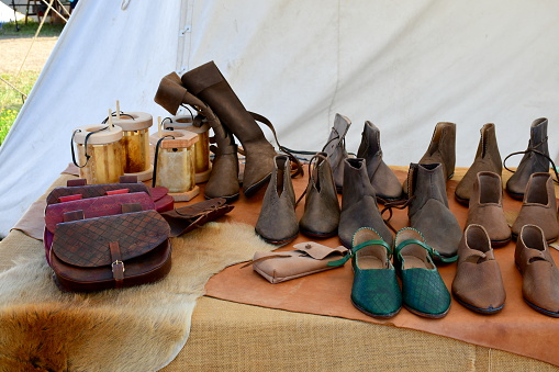 A view of a temporary stall or stand set up during a medieval fair or festival showing various types of shoes, boots, purses, mugs, lanterns and other everyday items seen on a sunny summer day
