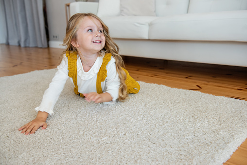Little curly blonde girl is lying on the carpet on the floor. Toddler is wearing a white shirt and a mustard yellow corduroy skirt. Carefree childhood. Happy holidays. Return to school. Copy space