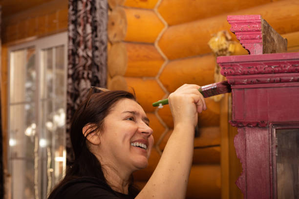 mulher feliz com sorriso pintando armário ornamentado de madeira em rosa com pincel com janela no fundo. reaproveitamento de coisas antigas antigas. sala de trabalho de renovação de móveis para casa - patination - fotografias e filmes do acervo