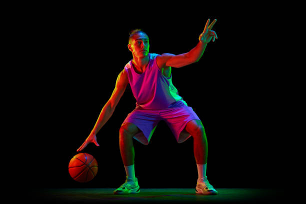 istock Game strategy. Young guy, basketball player in uniform dribbling ball against black studio background in neon light 1680015251
