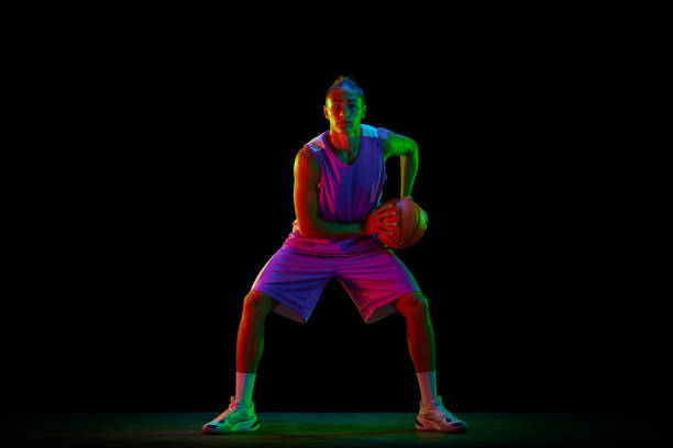 istock Young, active, motivated guy, basketball player in uniform standing in position with ball against black studio background in neon light 1680015280