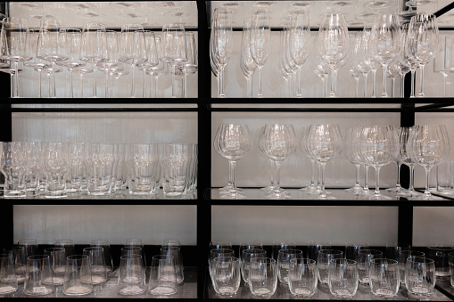 A wineglass storage rack displays an array of pristine, transparent glassware, including crystal-clear wine glasses. This elegant arrangement hints at a setting prepared for a sophisticated event or gathering. The immaculate rows of wine glasses evoke a sense of readiness and anticipation, perfectly organized and awaiting their moment of service. Each glass exudes an aura of elegance and fragility, embodying the gloss and refinement that accompanies fine dining and catering. Whether it's a banquet, a wine-tasting event, or a special occasion, this collection of glassware is a testament to the art of presentation and service. The background showcases the immaculate preparation of tableware, promising a dining experience marked by sophistication and attention to detail. It's an embodiment of the elegance and precision that elevate moments of celebration and indulgence.