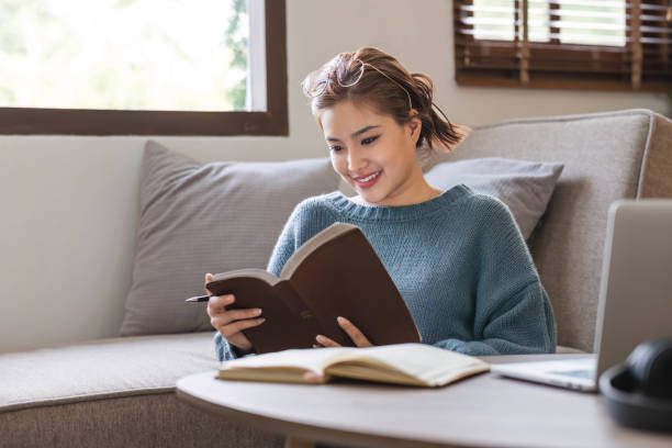 istock Attractive young Asian female reading a book while relaxing at living room. 1680021092