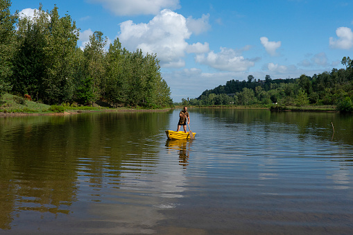 A young man standing and rowing with an oar on the calm lake on his yellow boat against a beautiful landscape in summer.