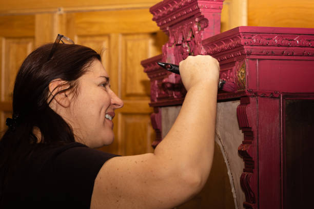 mulher branca sorridente colorindo armário ornamentado de madeira em vermelho com pincel grande com portas no fundo. reaproveitamento de coisas antigas antigas. sala de trabalho de renovação de móveis para casa - patination - fotografias e filmes do acervo