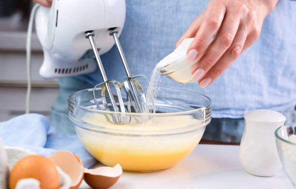 A woman beats eggs with a mixer and adds sugar while standing in the kitchen at home. Cooking and baking desserts at home. Selective focus.