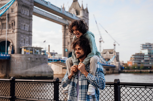 A mother takes photos of her husband and son  as they pose in front of some London landmarks with the son on his fathers shoulders.