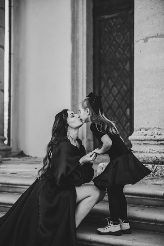 Mother kisses daughter stands on stairs near large columns of ancient temple at sunset. Child hold hands mom near Baroque Catholic church. Parent hugging happy kid on autumn day. Black and white photo