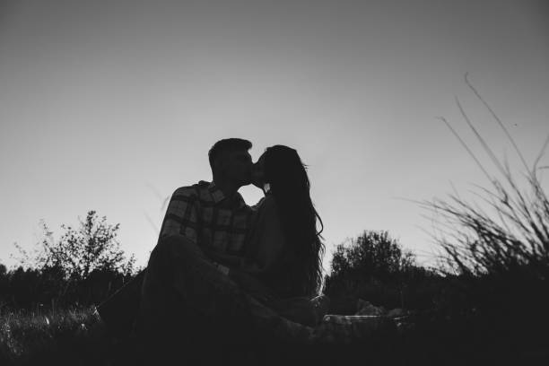 pareja sentada en una manta abrazándose en la hierba en el campo al atardecer. una joven feliz besa al hombre que pasa tiempo juntos en la naturaleza. concepto vacaciones en familia. hembra abraza macho en picnic. foto en blanco y negro - silhouette kissing park sunset fotografías e imágenes de stock