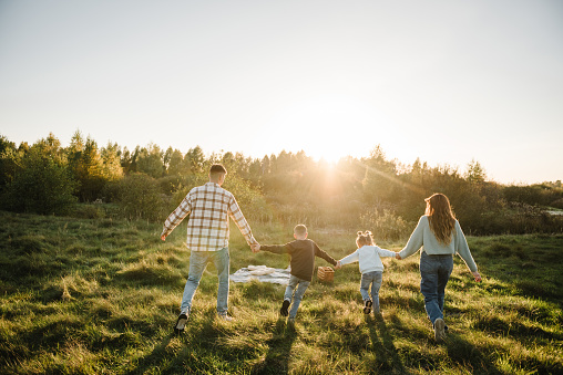 Mom, dad, daughter and son walk back in green grass in field. Happy young family with children spending time together, running outside, go in nature at sunset. Concept of family holiday outdoors.