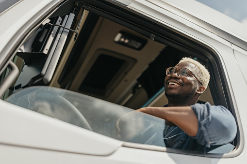 A cheerful African-American truck driver in his late 20s sits confidently in the driver's seat of his semi-truck show from the window. He is ready to embark on a successful journey in the trucking business.