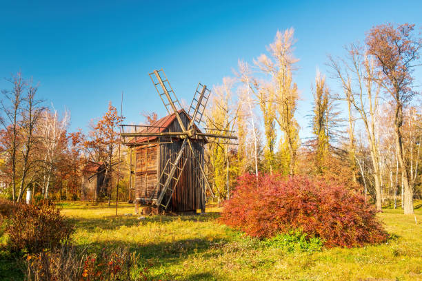 A picturesque autumn scene featuring a wooden windmill set in a rural landscape. The image captures the vibrant colors of fall foliage under a clear blue sky