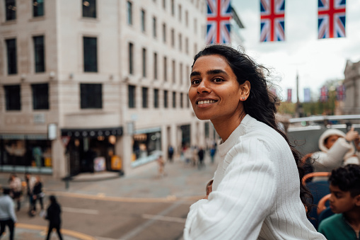 A woman leans on the rail of an open top bus and looks at the sights with a smile.