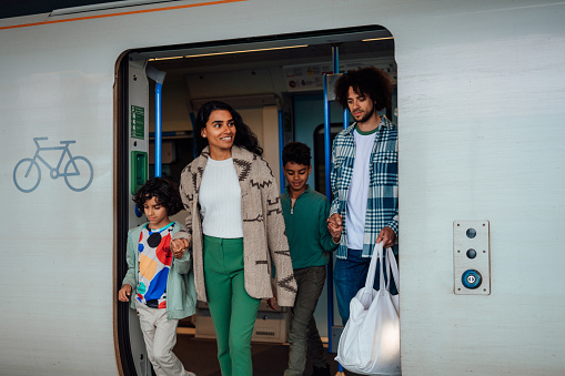 A young family hold hands and step off a train and onto a platform.