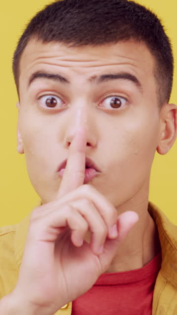 Secret, face and finger of man on lips in studio for sign, privacy and warning on yellow background. Portrait, model and quiet for gossip, whisper and noise for emoji, mystery and confidential news