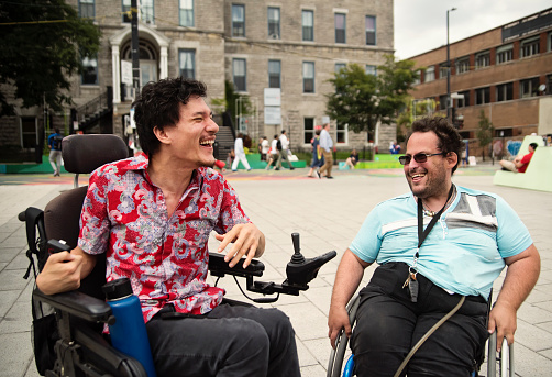 Friends living with disabilities meeting in city street. One is from indonesian ethnicity and a member of the LGBTQ community. The other is caucasian with sunglasses. Both are using wheelchairs. Horizontal waist up outdoors shot with copy space. This was taken in Montreal, Quebec, Canada.