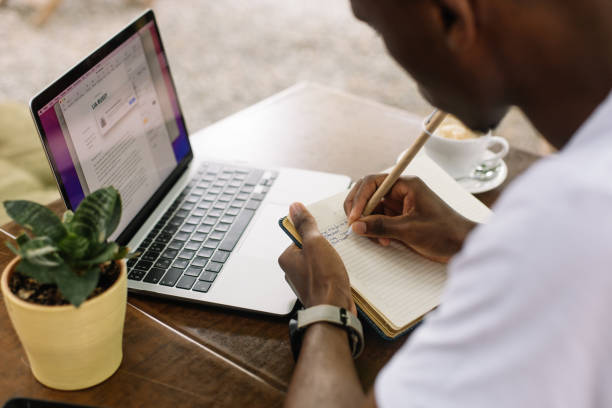 istock Rearview interracial man using laptop, writing notes in textbook and drinking coffee in cafe with wireless technology. 1680449976