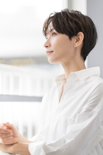 A Japanese woman is smiling by the window.She has short dark brown hair and wears a white shirt.