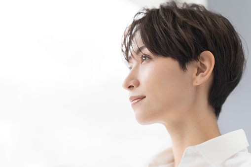 A Japanese woman is smiling by the window.She has short dark brown hair and wears a white shirt.